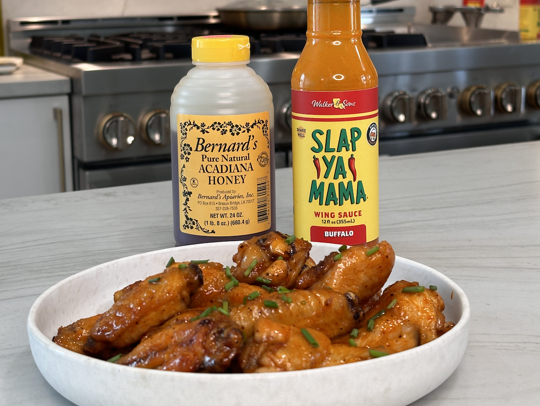 Plate of honey buffalo wings in a kitchen, with a bottle of honey and a bottle of Slap Ya Mama Wing Sauce placed behind the wings