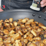 Image of breakfast potatoes in a pan, with a person holding a can of Garlic Salt Signature Blend seasoning over the pan