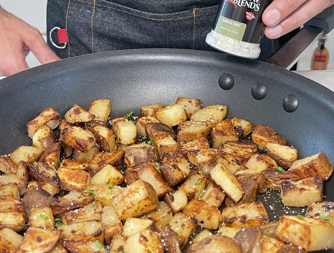 Image of breakfast potatoes in a pan, with a person holding a can of Garlic Salt Signature Blend seasoning over the pan